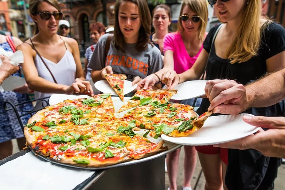 A group of people are serving themselves slices of a large freshly-baked pizza topped with cheese tomatoes and basil