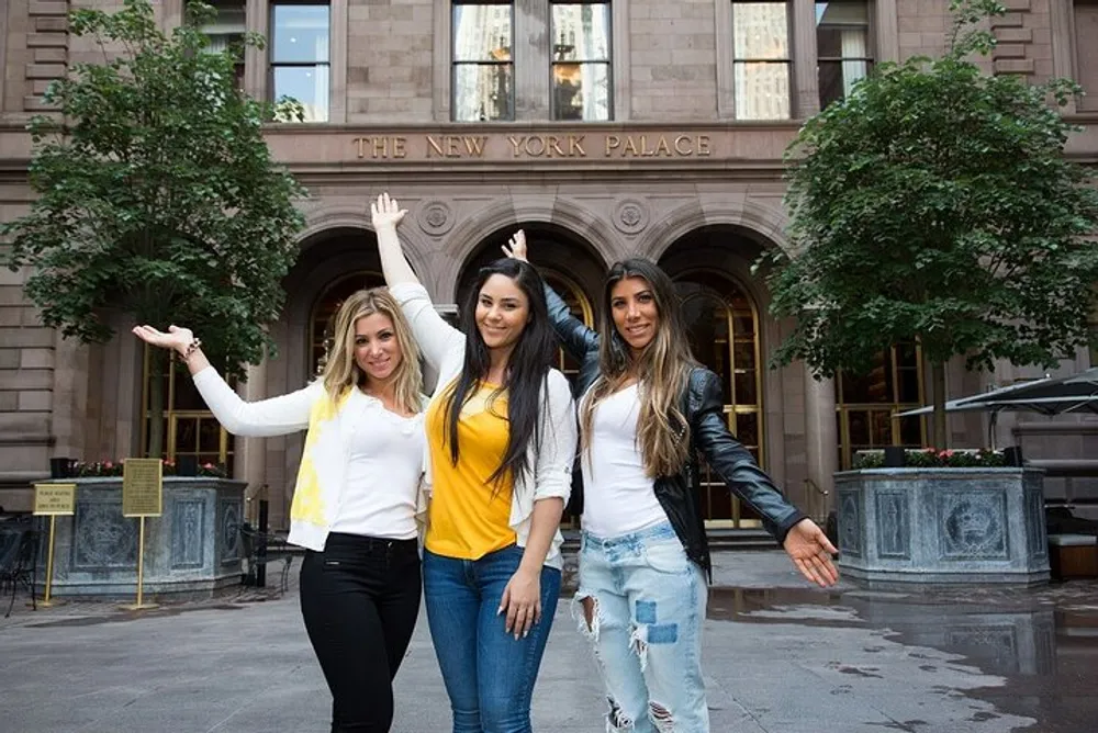 Three smiling women pose in front of the New York Palace Hotel with one arm extended upwards