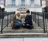 Two people are sitting on the stairs in front of the Museum of the City of New York smiling at the camera
