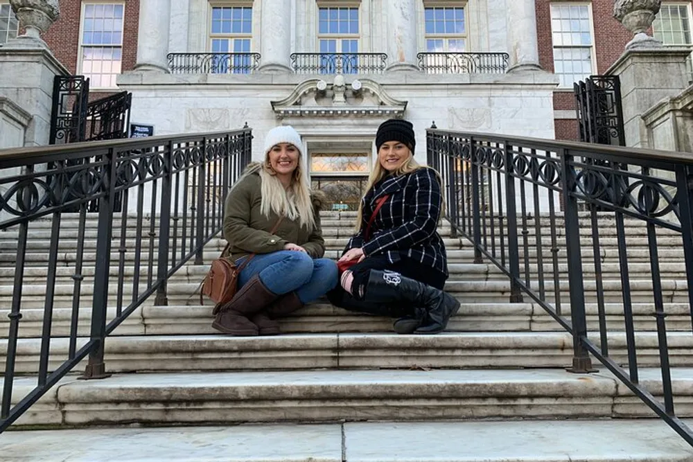 Two smiling individuals are sitting on the steps in front of an ornate building with classical architecture