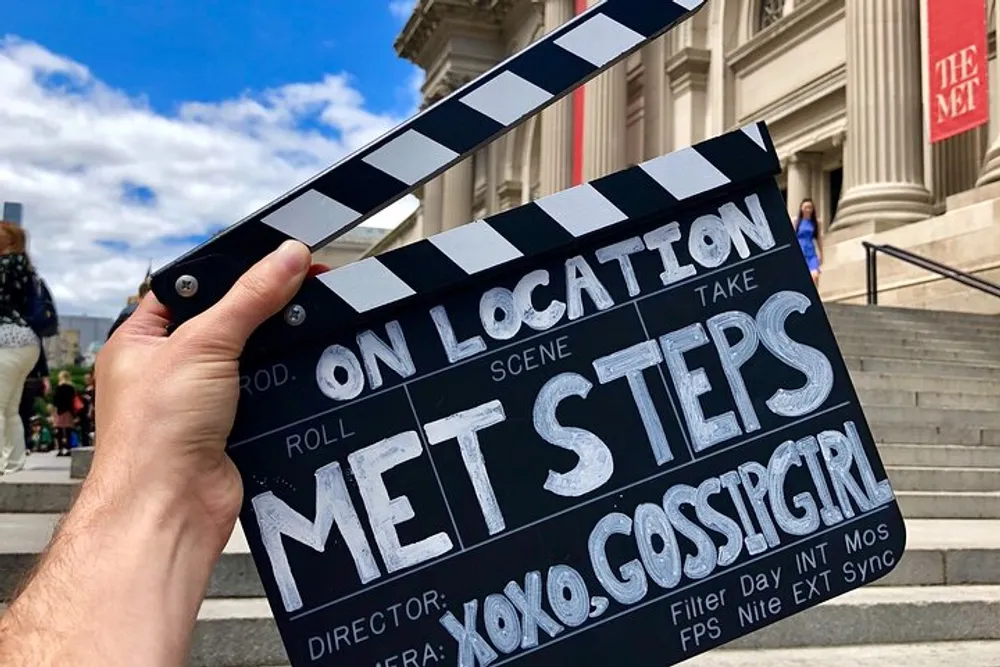 A person is holding a film clapperboard labeled MET STEPS XOXO GOSSIP GIRL in front of the stairs of the Metropolitan Museum of Art suggesting a filming location related to the television show Gossip Girl