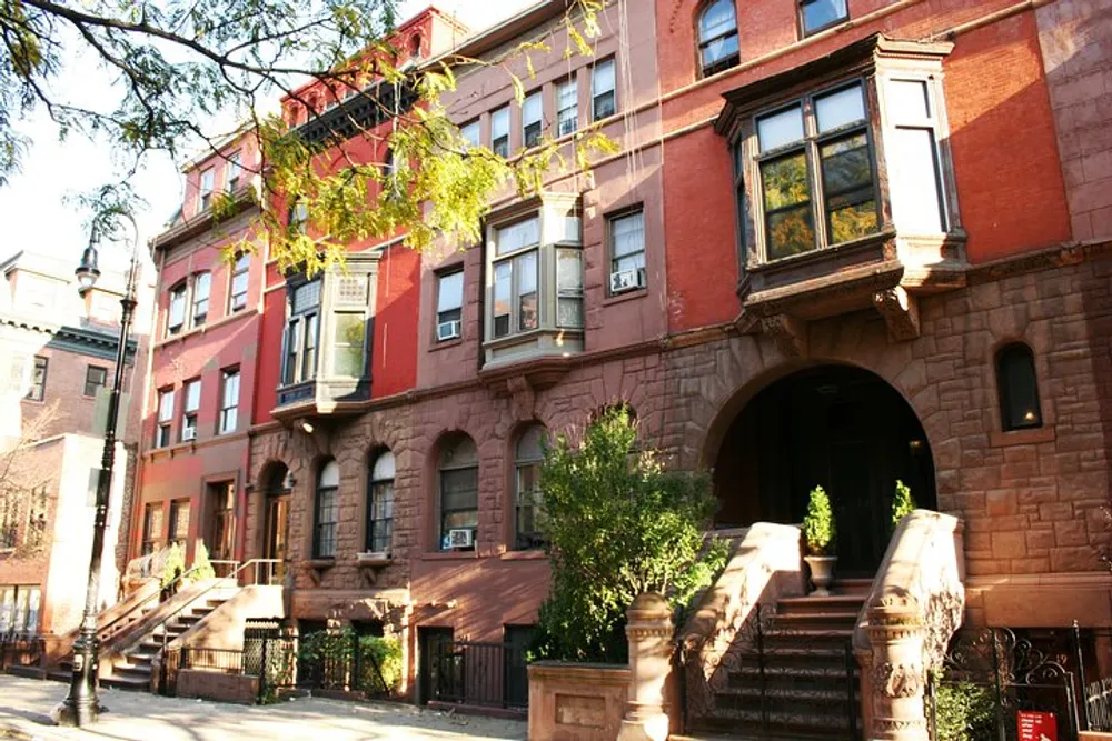 The image features a row of traditional brownstone townhouses basking in the sunlight typical of those found in many New York City neighborhoods