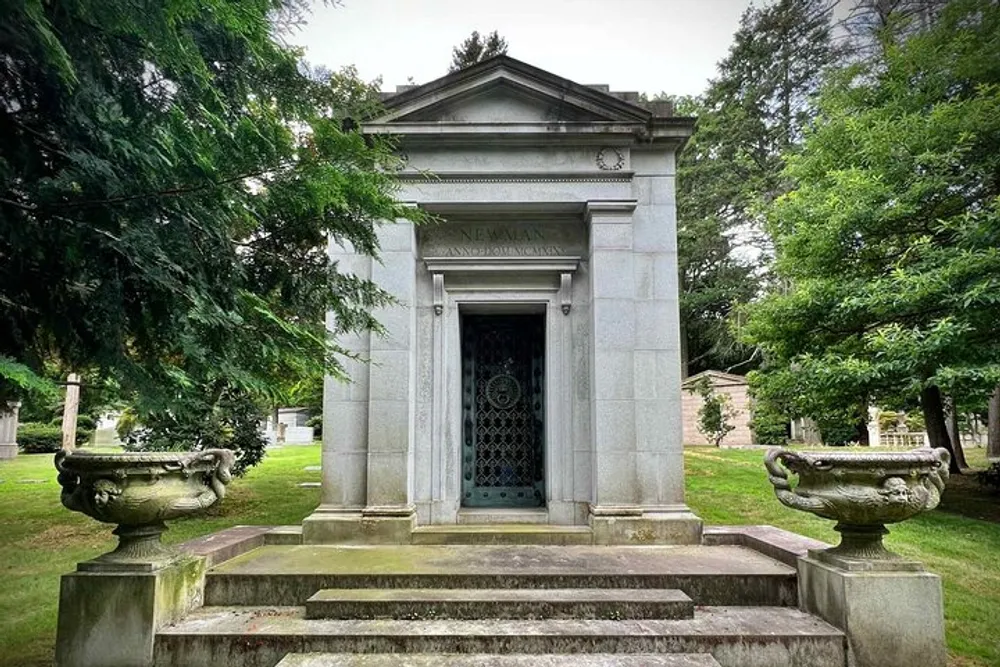 The image shows a stately vintage mausoleum with the name NEWMAN above the entrance ornate doors flanked by urns set in a tranquil cemetery
