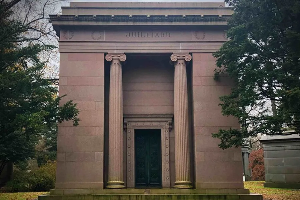 The photo depicts a stately mausoleum with the name Julliard engraved at the top featuring classic architectural elements such as tall columns and an ornate bronze door