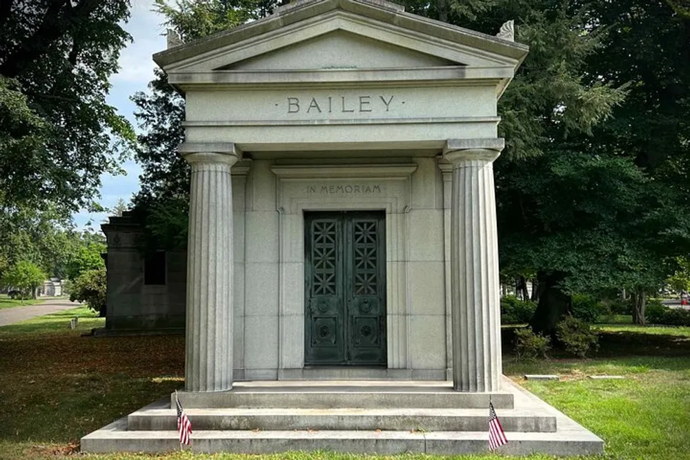 The image depicts the front view of a grand neoclassical mausoleum engraved with the name BAILEY and the phrase IN MEMORIAM adorned with two American flags positioned symmetrically at its base