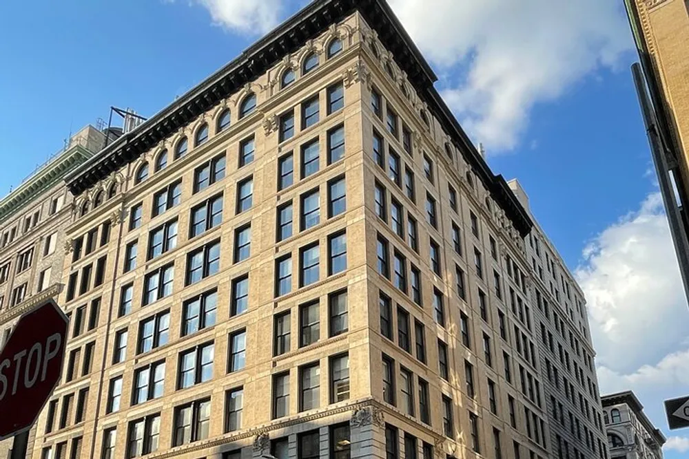 The image shows a large multi-story corner building with classic architectural details under a blue sky with a stop sign in the foreground