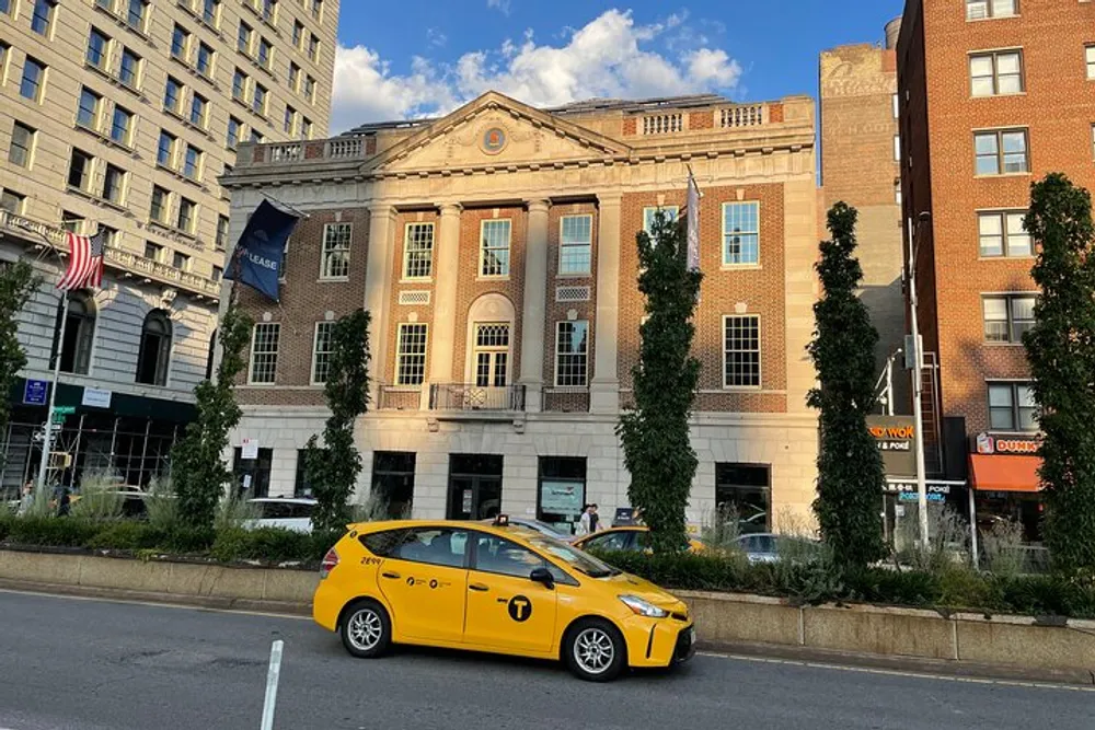A yellow taxi drives past a row of buildings with an American flag and tall green trees on a sunny day