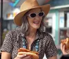 A smiling person wearing a sun hat and sunglasses is having a meal and a pleasant conversation at an outdoor dining area