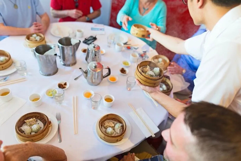 A waiter is serving dim sum to a group of people at a restaurant table covered with various foods and teaware