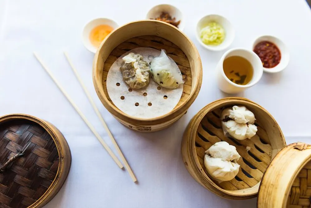 The image shows a dim sum meal with dumplings in bamboo steamers accompanied by chopsticks and various condiments on a white tablecloth