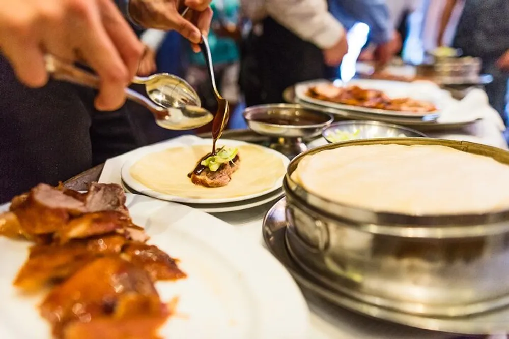 A person is drizzling sauce on a Peking duck wrap in a lively dining setting