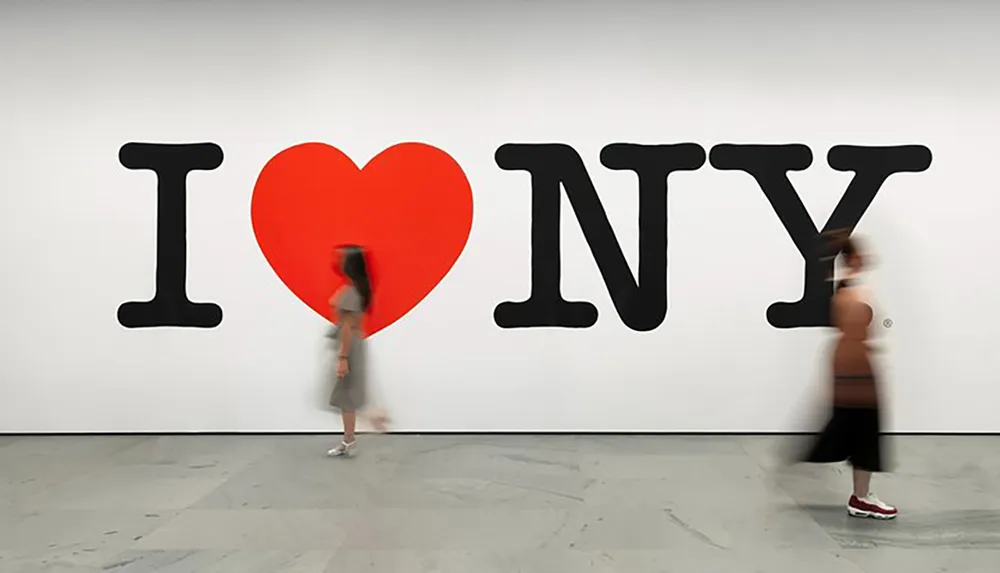 Two people are walking past the iconic I Love NY slogan displayed prominently on a gallery wall