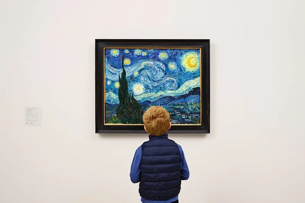 A child is standing in front of and observing a framed painting of a starry night over a small town