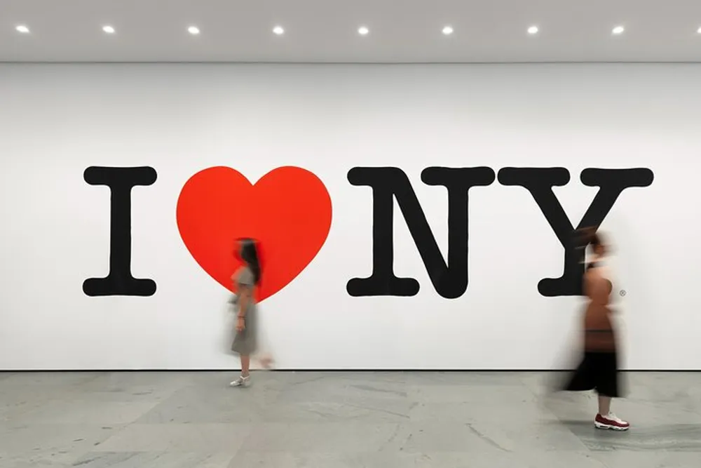The image shows a large I Love NY mural on a wall with two people walking past one blurred indicating movement