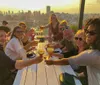 A group of people are enjoying drinks and toasting at an outdoor table with a view of a city skyline bathed in the warm glow of sunset