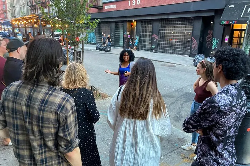 A group of people stands on a city sidewalk attentively listening to a woman who is speaking and gesturing with her hands