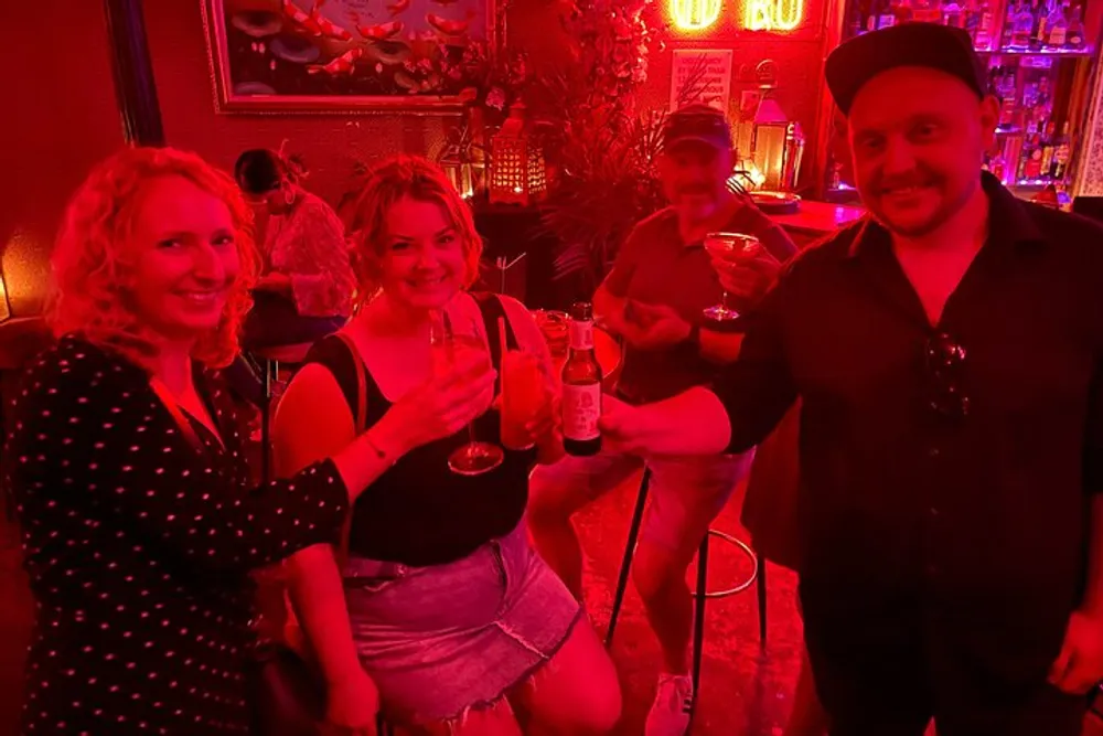 Three people are smiling and toasting drinks in a dimly lit bar with a vibrant red ambiance
