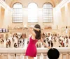 A woman in a pink dress is seen from behind holding a hat and looking over the bustling interior of a grand train station