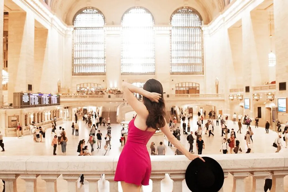 A woman in a pink dress is seen from behind holding a hat and looking over the bustling interior of a grand train station