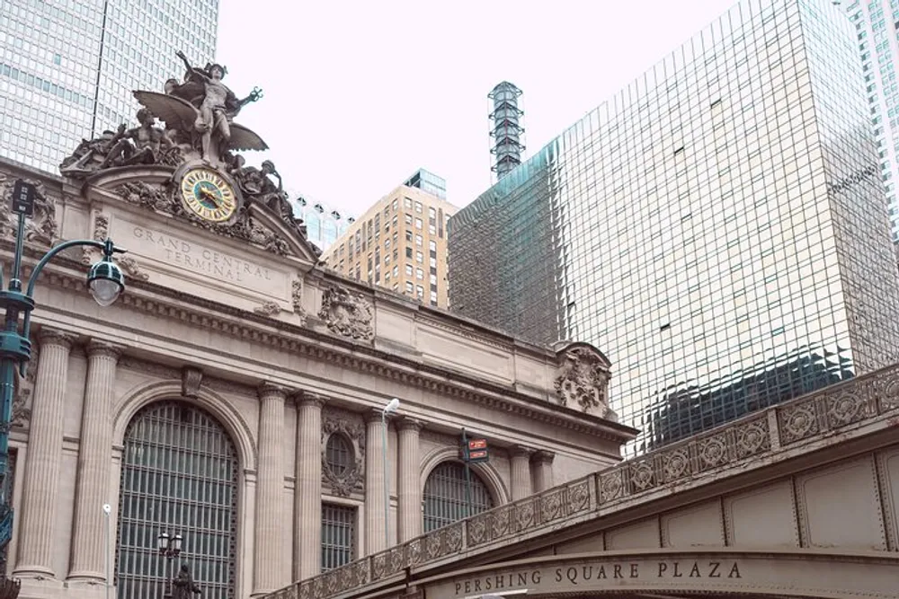The image shows a section of the ornate facade of Grand Central Terminal with its sculpture and clock set against a backdrop of modern skyscrapers in an urban setting