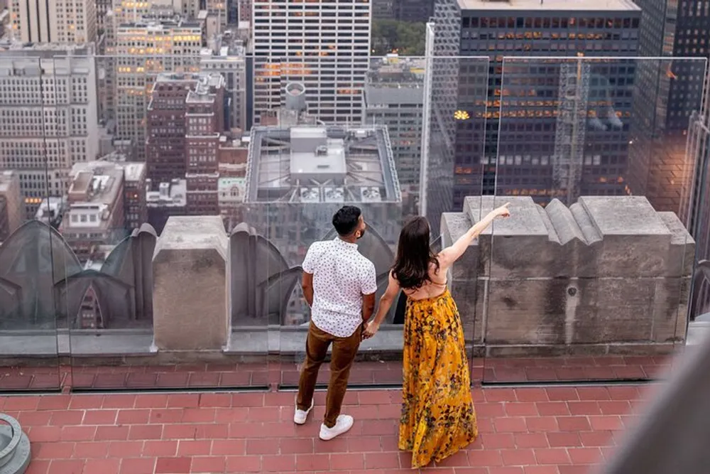 A man and a woman are standing on a high vantage point overlooking a cityscape with the woman pointing at something in the distance
