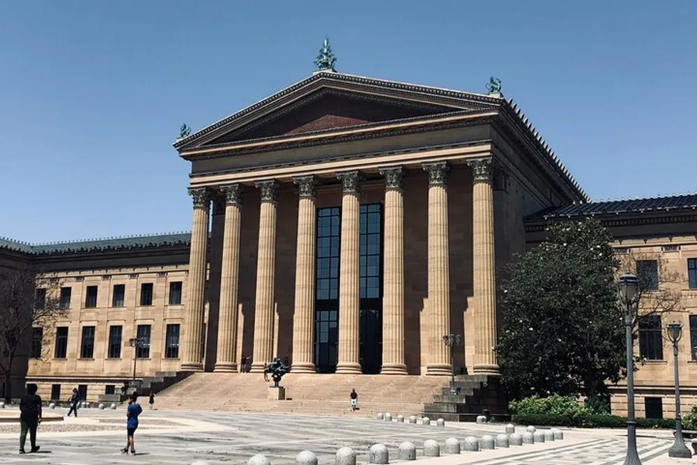 The image shows a grand neoclassical building with tall columns and a wide staircase under a clear blue sky with people walking in front