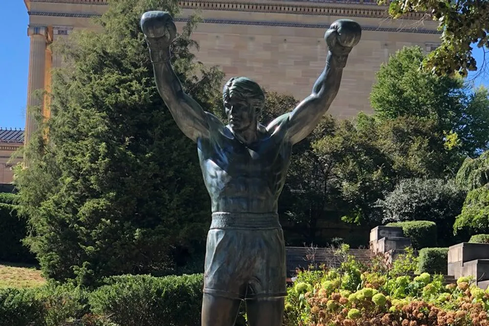 The image features a bronze statue of a boxer with his arms raised in victory positioned in front of a building with classical architecture amidst greenery