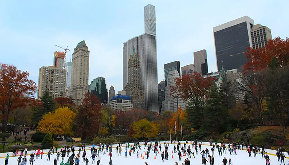 People are ice skating on an outdoor rink surrounded by the colorful foliage of a city park with a backdrop of towering skyscrapers