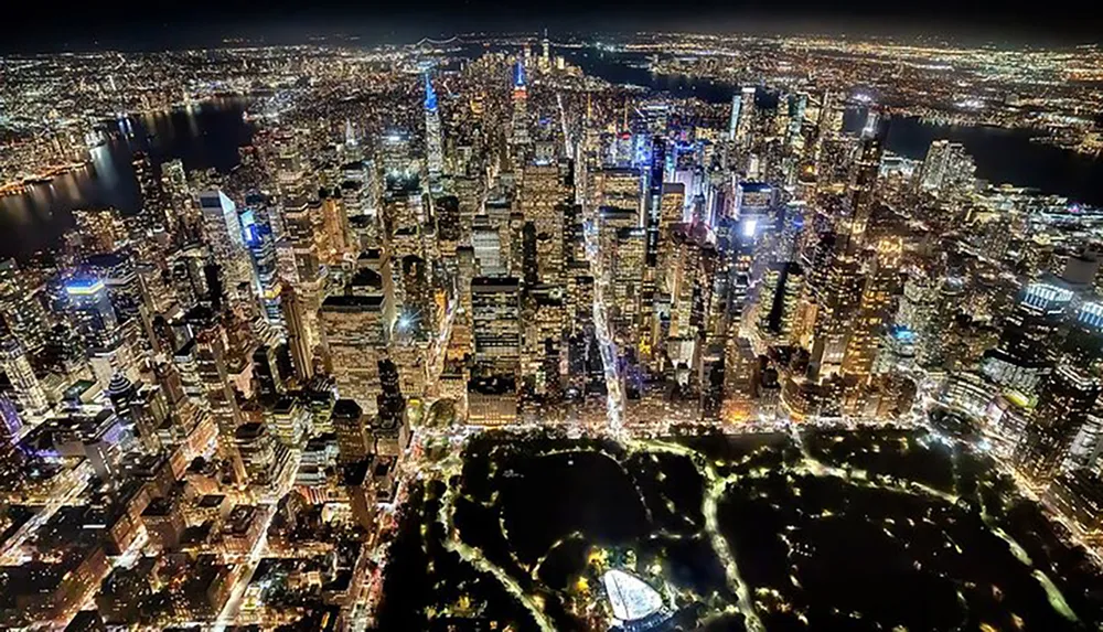 A nighttime aerial view showcases the glittering skyscrapers and dense urban landscape of Manhattan with Central Park providing a dark green respite amidst the city lights