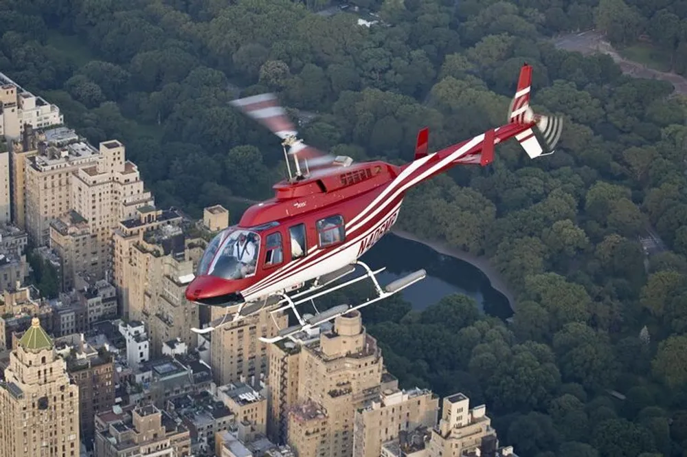 A red and white helicopter is flying over an urban area adjacent to a large park-like green space