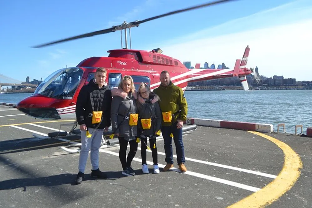 Four people are smiling for a photo in front of a red helicopter on a helipad near a body of water with a city skyline in the background