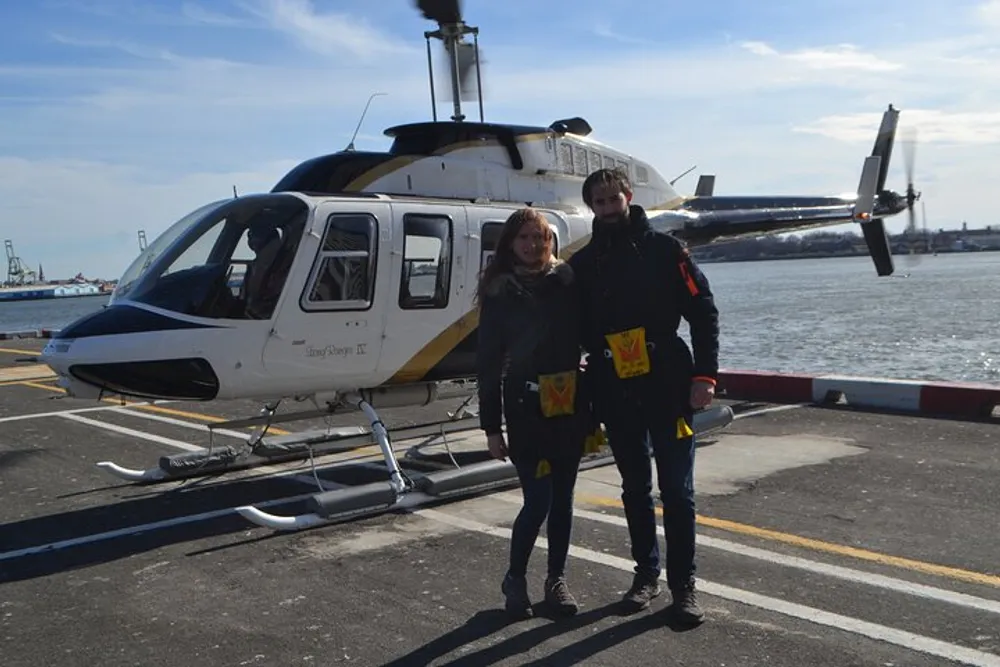 A man and a woman are smiling for a picture in front of a helicopter on a sunny day likely after or before a helicopter ride