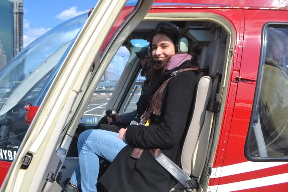 A person is smiling while seated inside a red helicopter wearing a headset and looking ready for flight