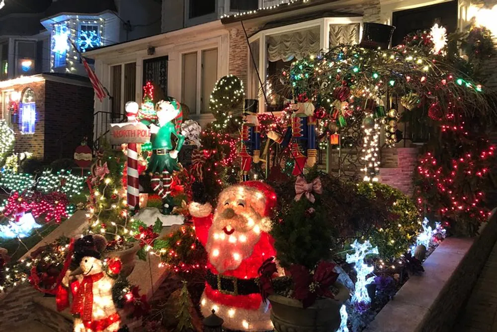 The image shows a festive outdoor Christmas decoration with illuminated figures including Santa Claus an elf and snowmen as well as lights and greenery adorning the house and walkway