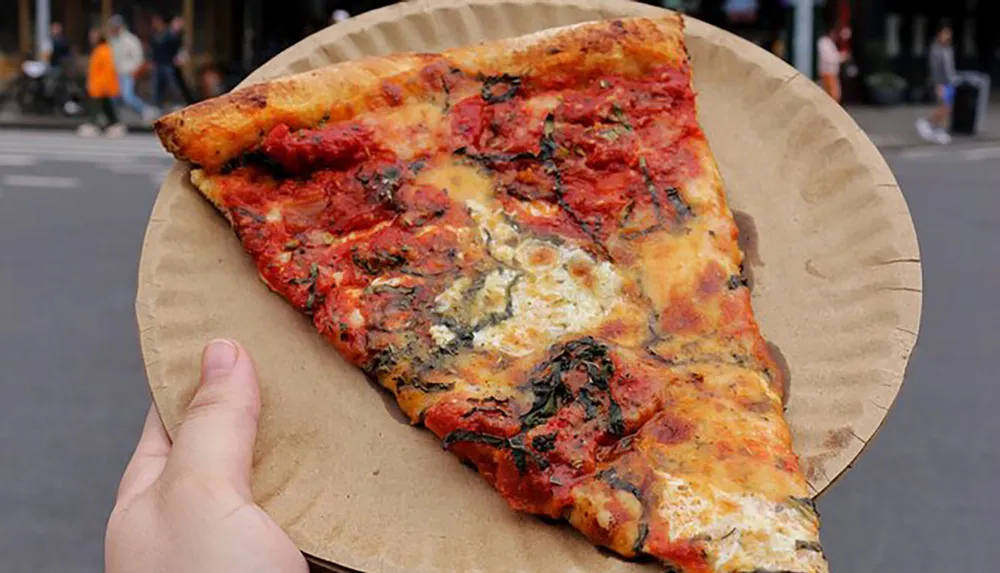 A person is holding a large slice of pizza on a paper plate in an urban setting