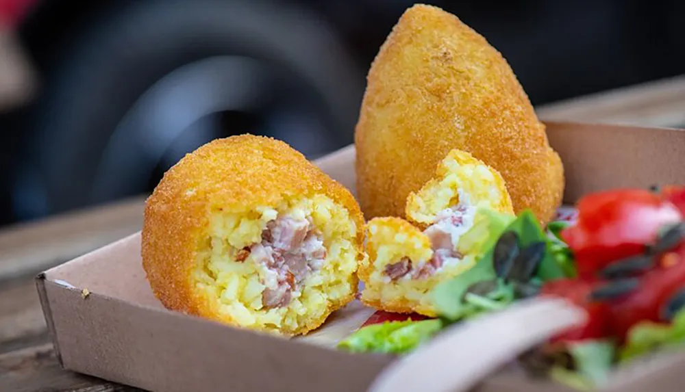 The image showcases two golden-brown arancini rice balls one of which is cut open to reveal a creamy risotto with ham filling served in a cardboard tray accompanied by a side salad