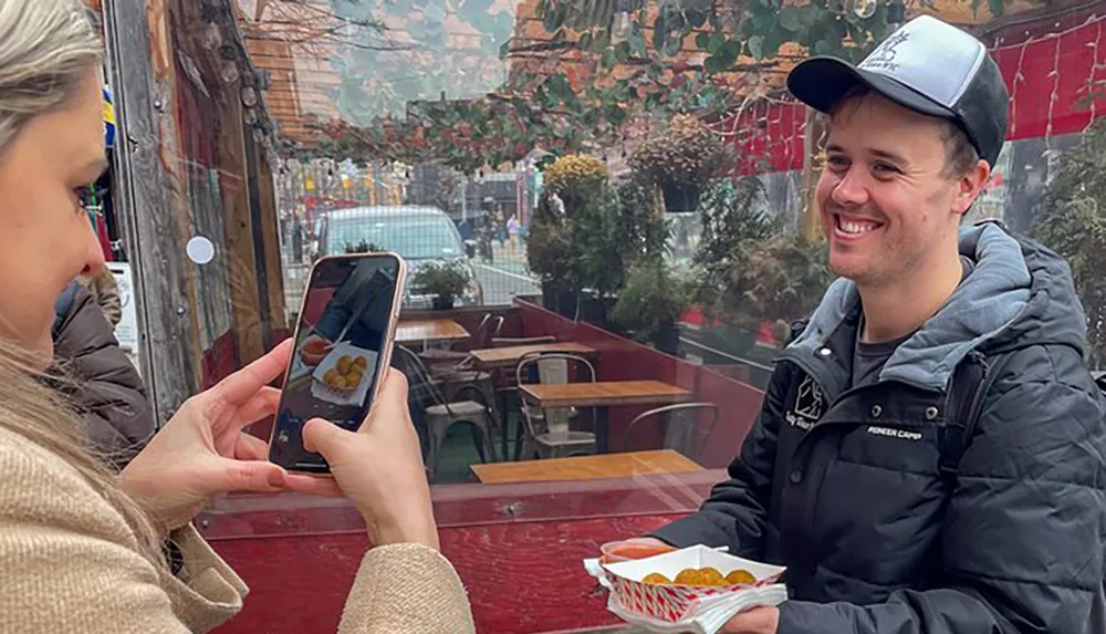 A person takes a photo of another person who is smiling and holding a tray of street food at an outdoor dining area