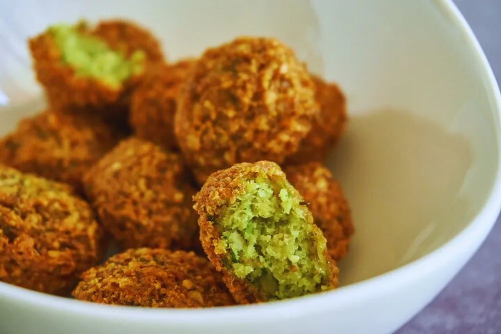 A bowl full of golden-brown falafel balls with one split open to show the green herby interior