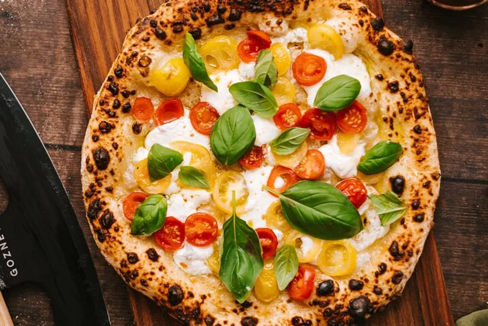 The image shows a freshly baked pizza adorned with melted cheese colorful cherry tomatoes and fresh basil leaves presented on a wooden table surface