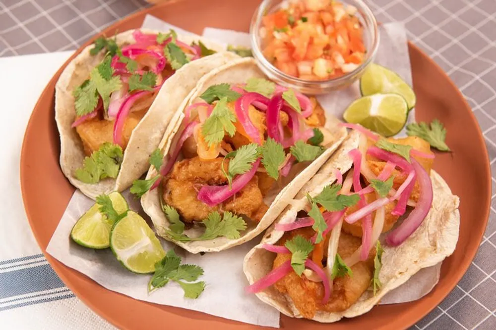 The image shows a plate of three fish tacos garnished with fresh cilantro and pickled red onions accompanied by lime wedges and salsa on the side