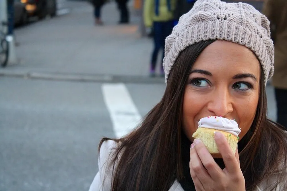 A woman wearing a knitted beanie is smiling as she is about to take a bite from a frosted cupcake outdoors