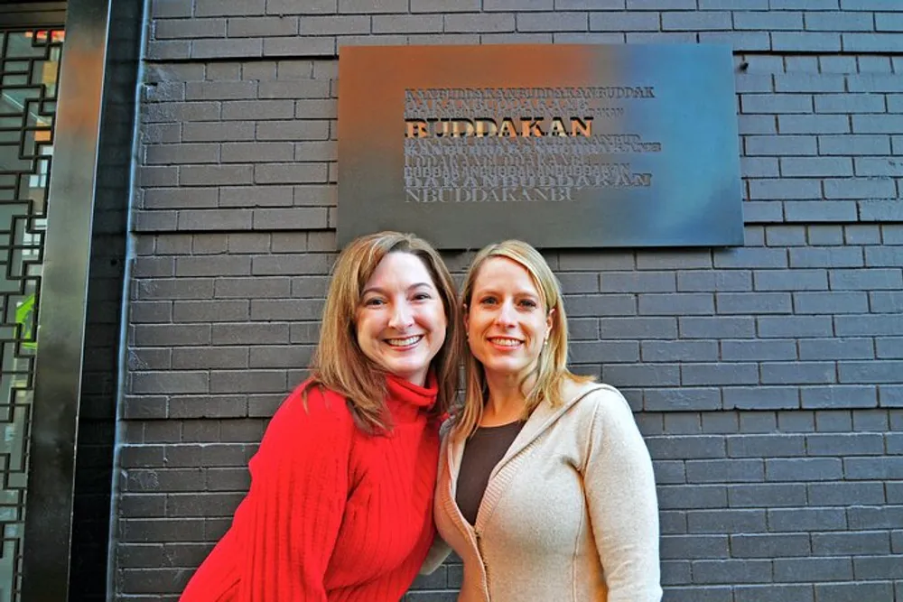 Two women are smiling at the camera in front of a dark brick wall that bears a metallic sign with indistinct text on it