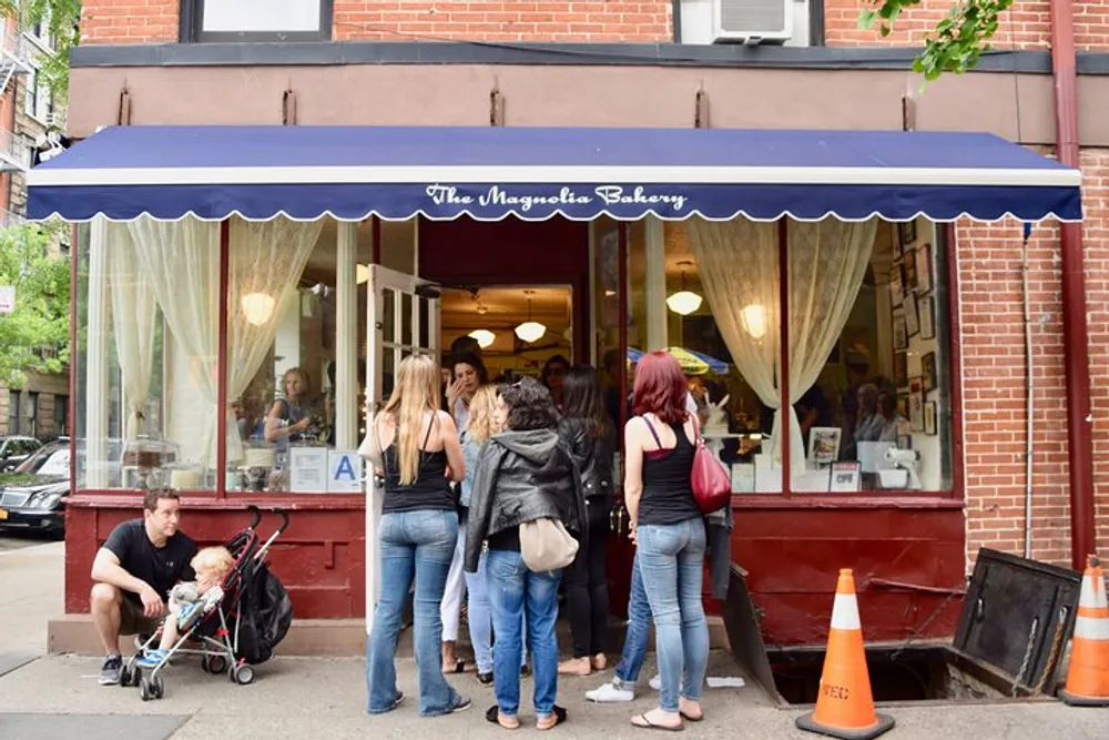 Customers stand in line outside The Magnolia Bakery which features a navy blue awning and classic storefront giving a glimpse into a busy and popular urban bakery scene