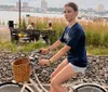 A person is riding a bicycle with a basket along a waterfront path with other park-goers and an urban skyline in the background