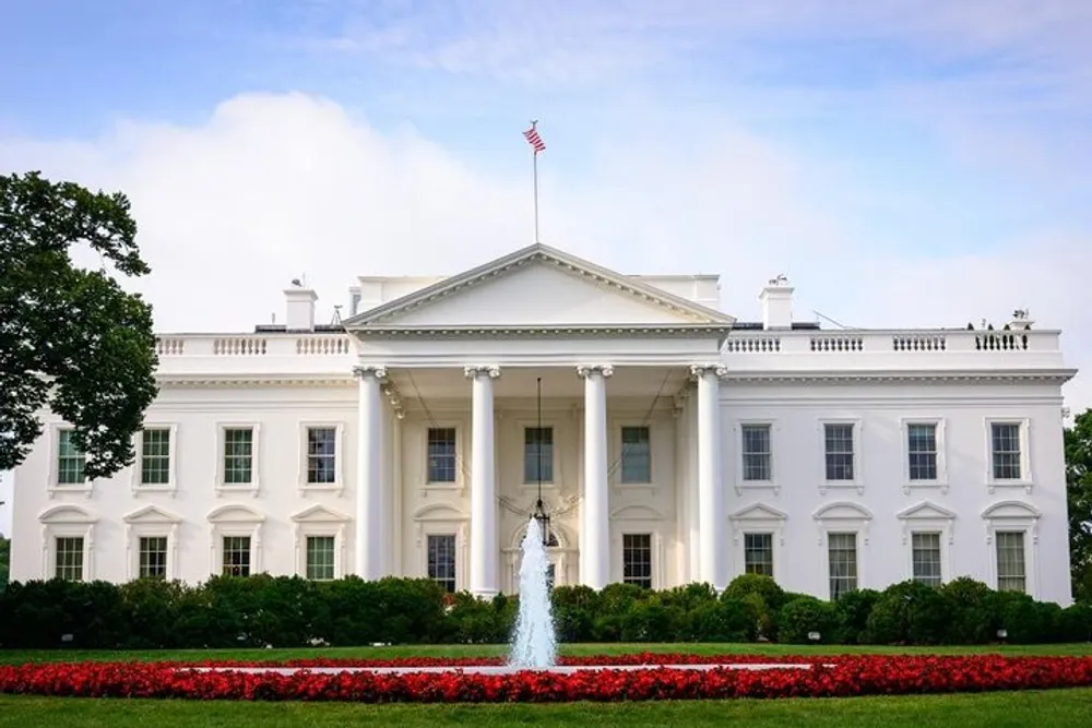 The image displays the White House the official residence and workplace of the President of the United States with its iconic neoclassical faade a central fountain and vibrant red flowers in the foreground