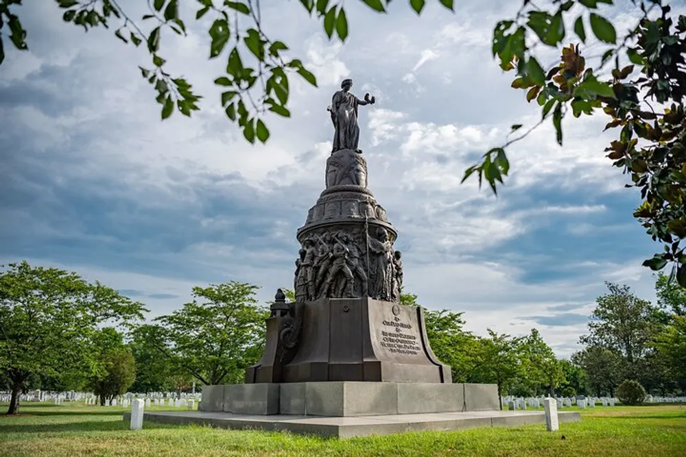 This image features an elaborate war memorial statue framed by greenery with a cloudy sky overhead and a multitude of grave markers spread out in the background