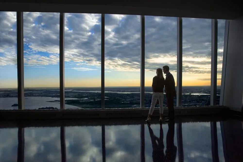 Two people are standing by wide windows enjoying a panoramic view of the sky at dusk with their silhouettes reflected on the shiny floor