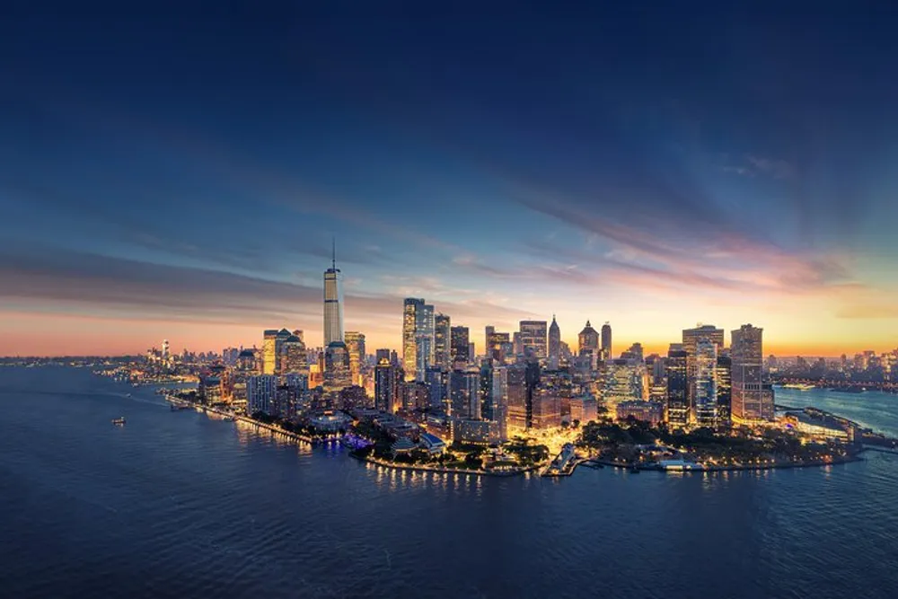 The image captures a stunning sunset view of a dense city skyline with skyscrapers illuminated against the fading light reflecting off the adjacent body of water