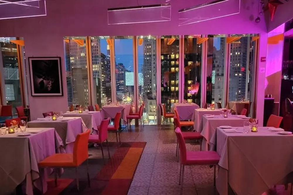 The image shows an upscale restaurant interior at twilight with neatly arranged tables and a panoramic view of a brightly-lit cityscape