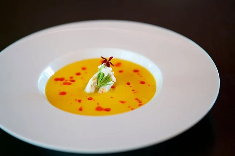 A bowl of smooth yellow soup elegantly plated with a dollop of cream and red oil droplets garnished with a delicate herb or flower